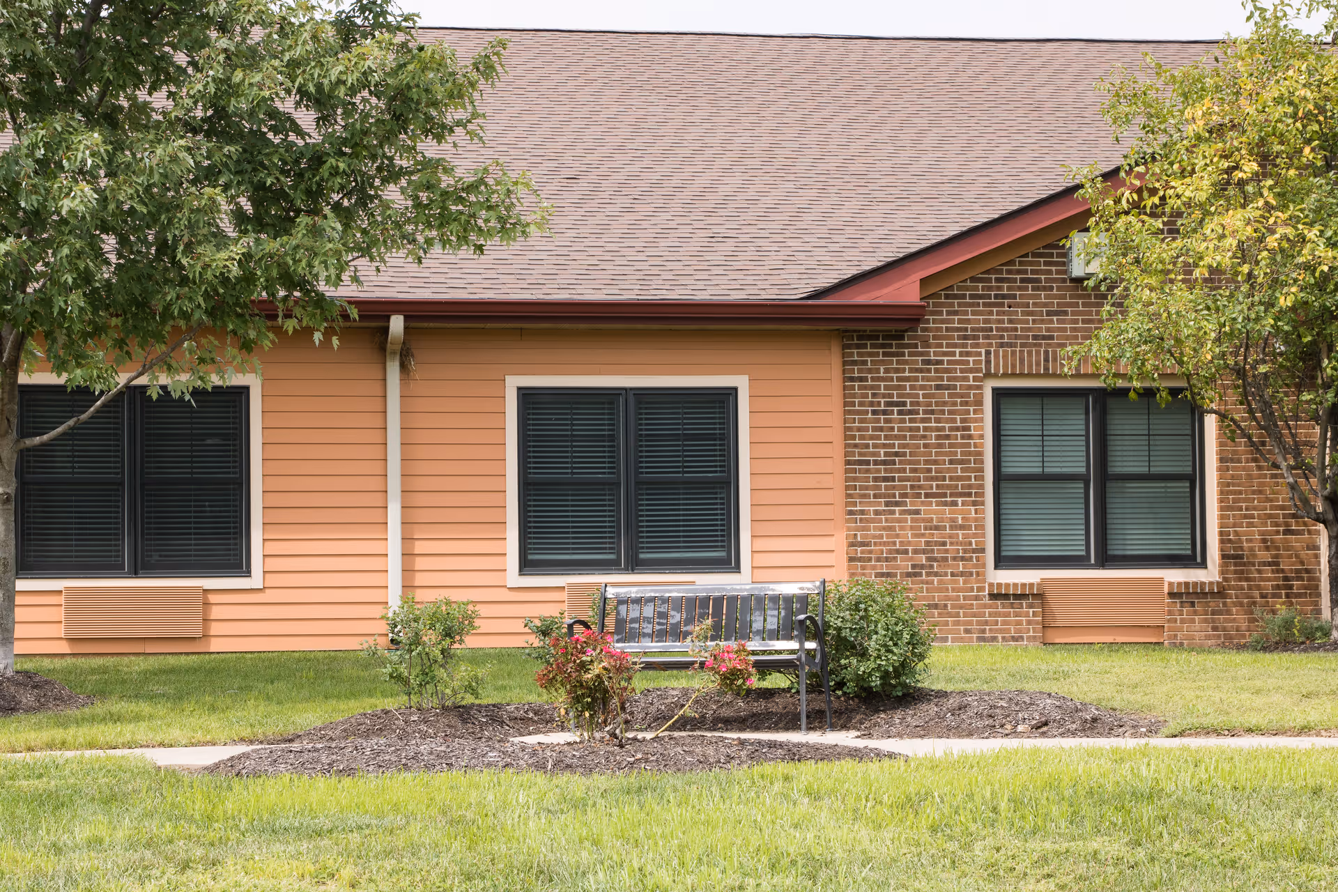 Outdoor view of a senior living facility building with a mix of orange siding and brick exterior. There are two windows with closed blinds, a bench in front of the building, small bushes, and trees on a well-maintained lawn.
