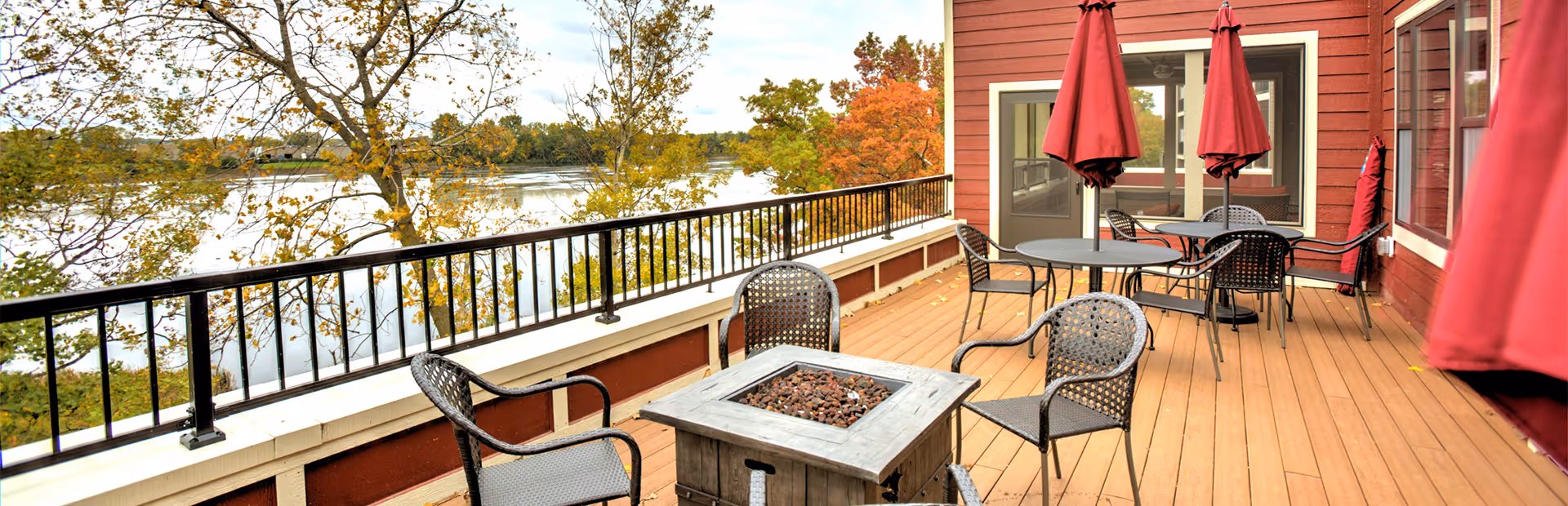 Outdoor patio area with several black metal chairs and tables, some tables have closed red umbrellas. The patio overlooks a body of water with trees showing autumn foliage in the background. The building exterior is red with white trim.