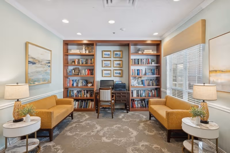 A cozy reading and work area in a senior living facility featuring two mustard yellow sofas facing each other with small round side tables and lamps beside each. Between the sofas is a built-in wooden bookshelf filled with books and decorative items, with a desk and chair in the center holding a computer and printer. The room has light-colored walls, a patterned carpet, and large windows with blinds on the right side.