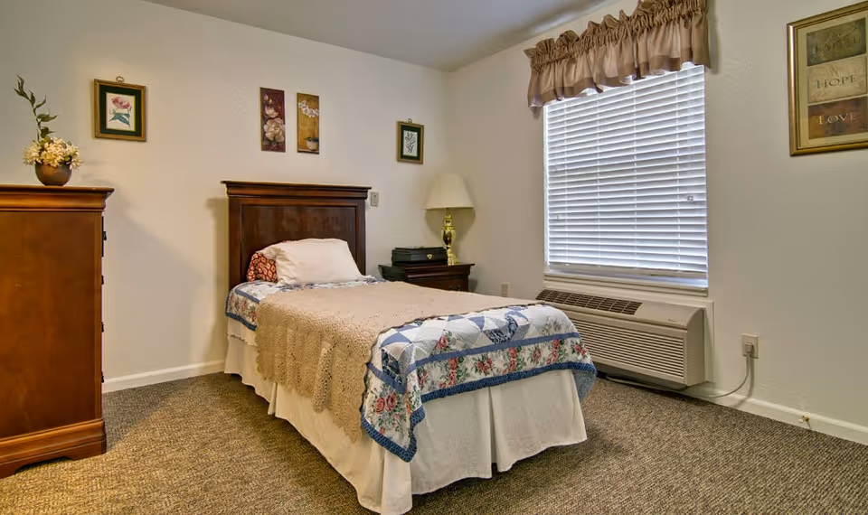 A small furnished bedroom with a single bed, wooden headboard and dresser, nightstand with lamp, and a window with blinds.