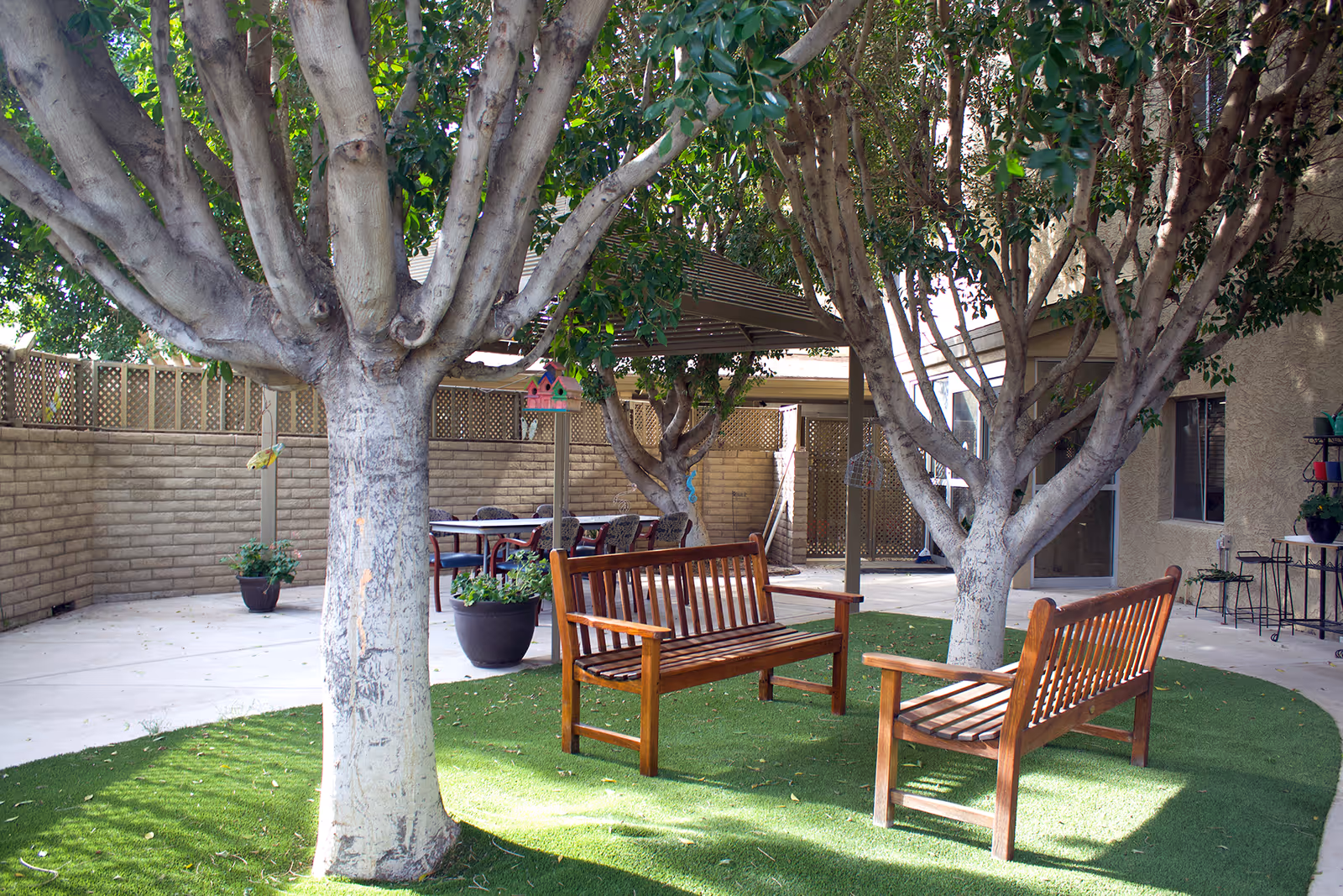 Outdoor courtyard area with two wooden benches placed on artificial grass under large trees. In the background, there is a patio with a table and chairs, potted plants, and a brick wall fence surrounding the space.
