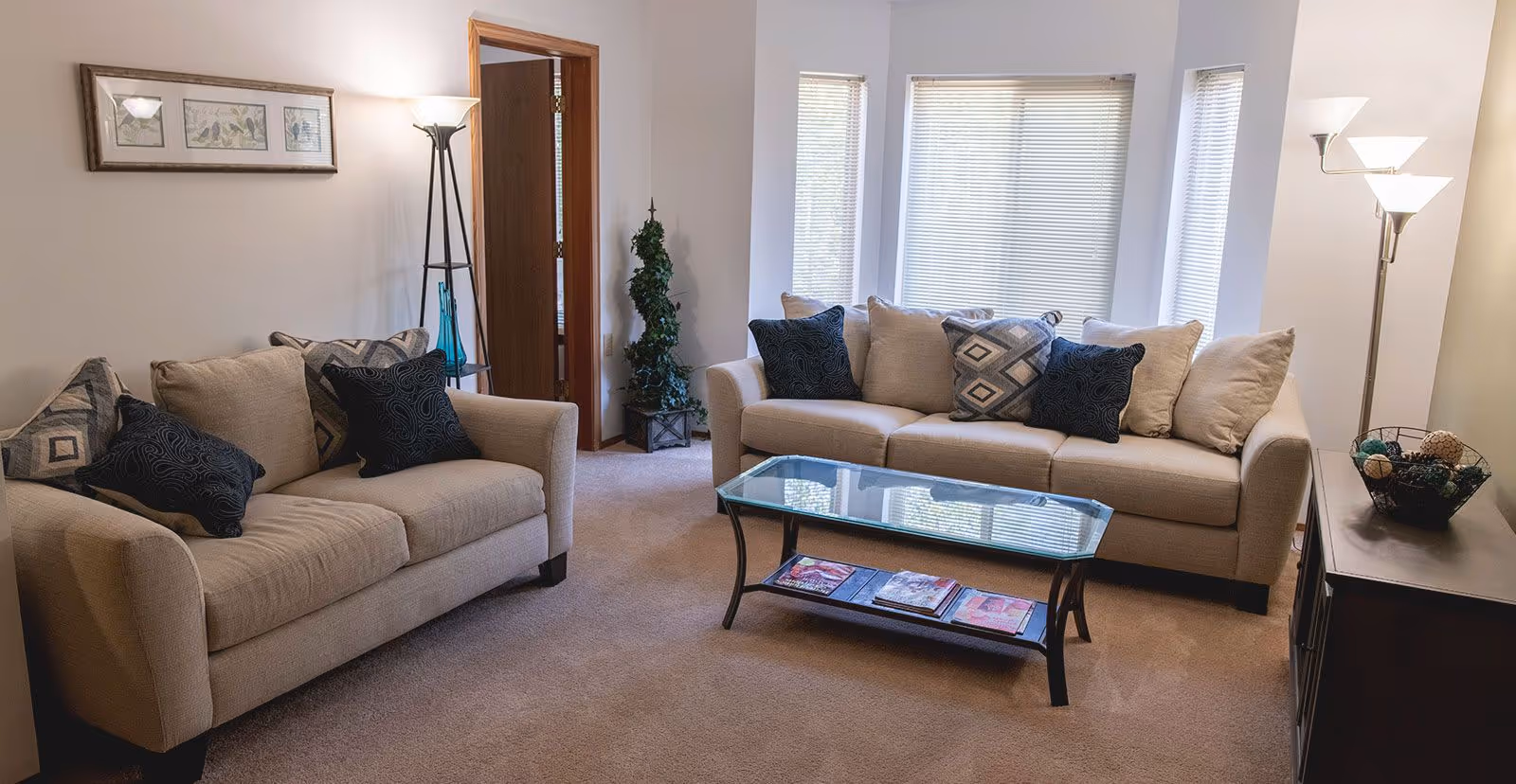 A bright living room with two beige sofas, a glass coffee table, decorative pillows, floor lamps, and a bay window.