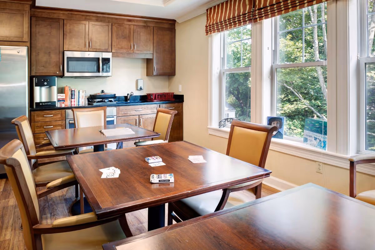 A bright room with wooden tables and chairs arranged for playing cards. The tables have playing cards and card boxes on them. The room has large windows with a view of green trees outside. In the background, there is a kitchen area with wooden cabinets, a microwave, a refrigerator, and a coffee machine.