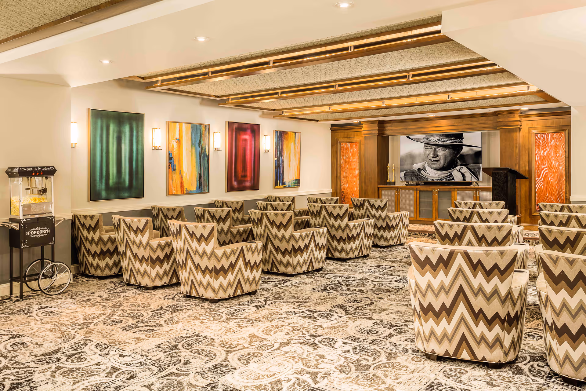 A carpeted interior screening room with chevron-patterned chairs facing a large TV screen, colorful wall art, and a popcorn cart.