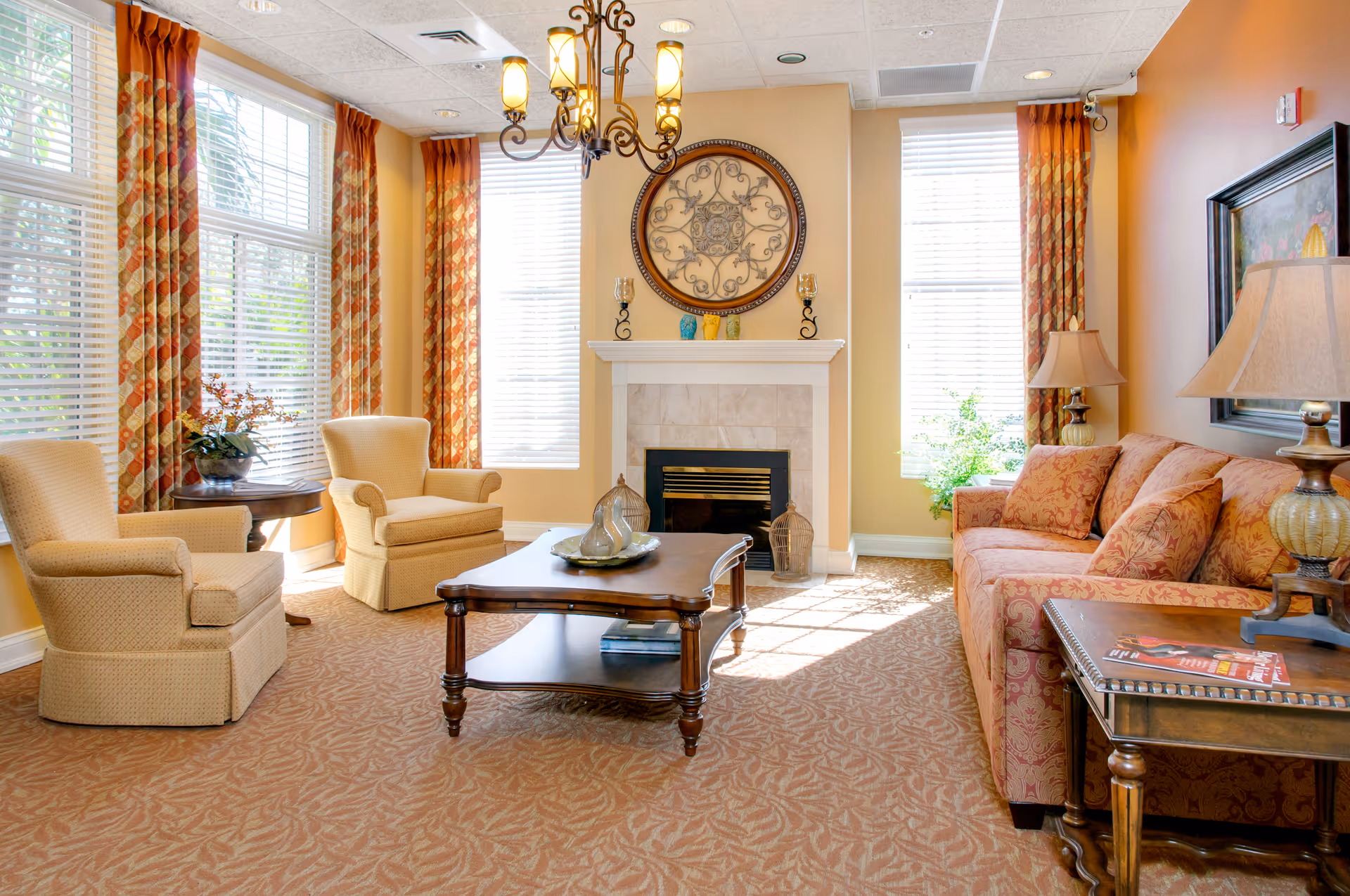 A bright and cozy living room with large windows covered by patterned orange curtains. The room features a beige armchair and loveseat, a wooden coffee table with decorative items, a fireplace with a round ornamental wall hanging above it, and a side table with a lamp and magazines. The walls are painted a warm yellow, and the carpet has a subtle leaf pattern.