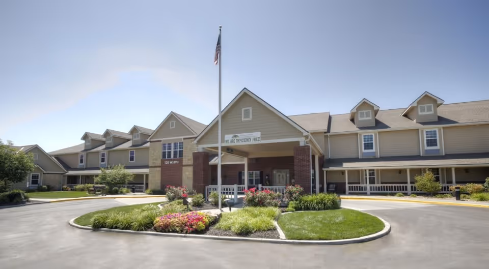 Front exterior view of a senior living facility named Vintage Park at Tonganoxie, featuring a two-story building with beige siding and brick accents, a covered entrance with a flagpole in front, and landscaped greenery and flowers around the driveway.