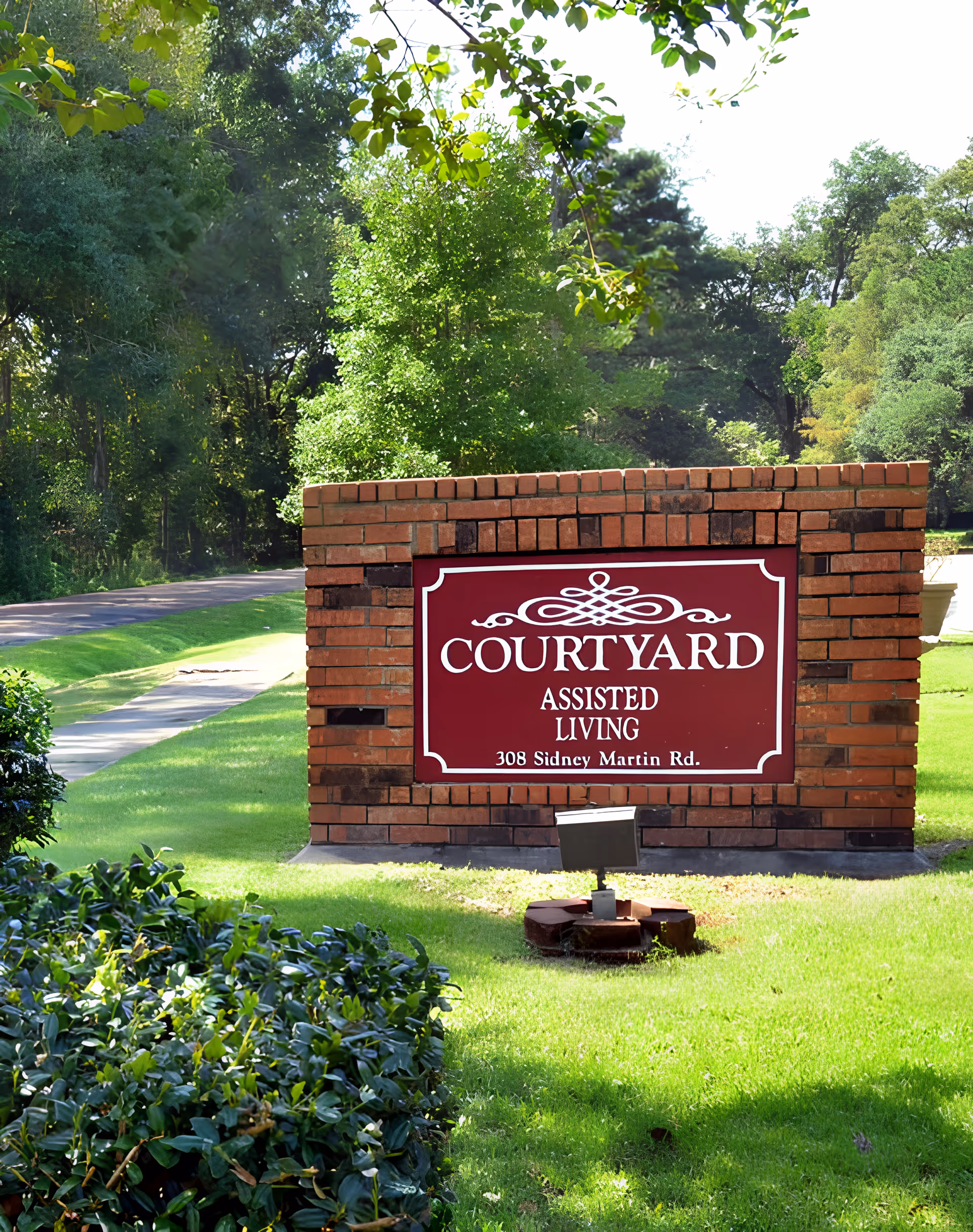 A brick sign for Courtyard Assisted Living with the address 308 Sidney Martin Rd., surrounded by green grass, bushes, and trees on a sunny day.