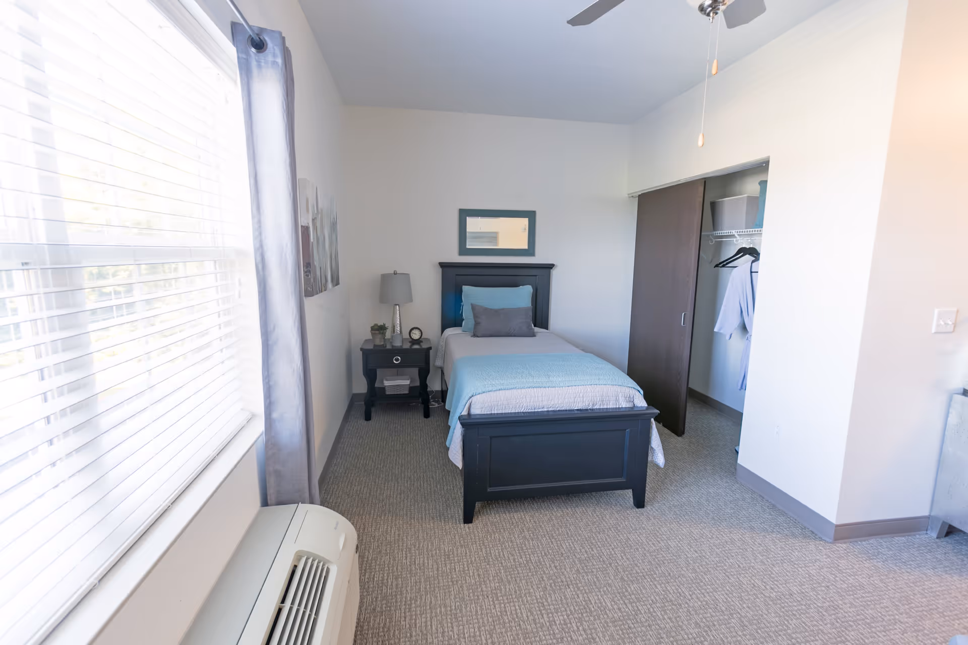 Bright, small bedroom with a single bed, nightstand, open closet and a large window with blinds.