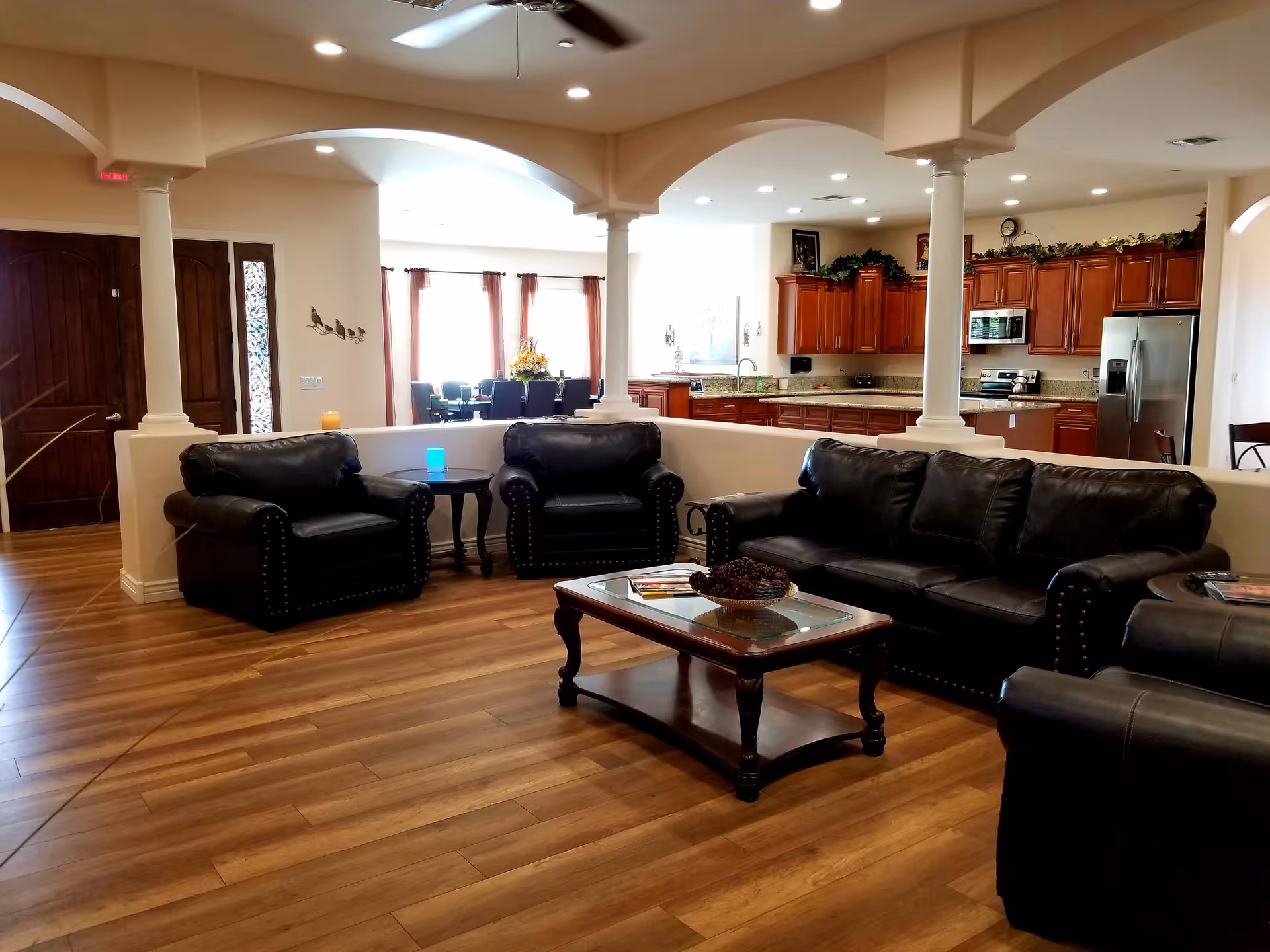 Spacious living room area with dark leather sofas and armchairs arranged around a wooden coffee table with a decorative bowl. The room has wooden flooring, white walls, and is open to a kitchen area with wooden cabinets, granite countertops, and stainless steel appliances. There is a dining area with a table and chairs near large windows with curtains, and ceiling fans with lights overhead.