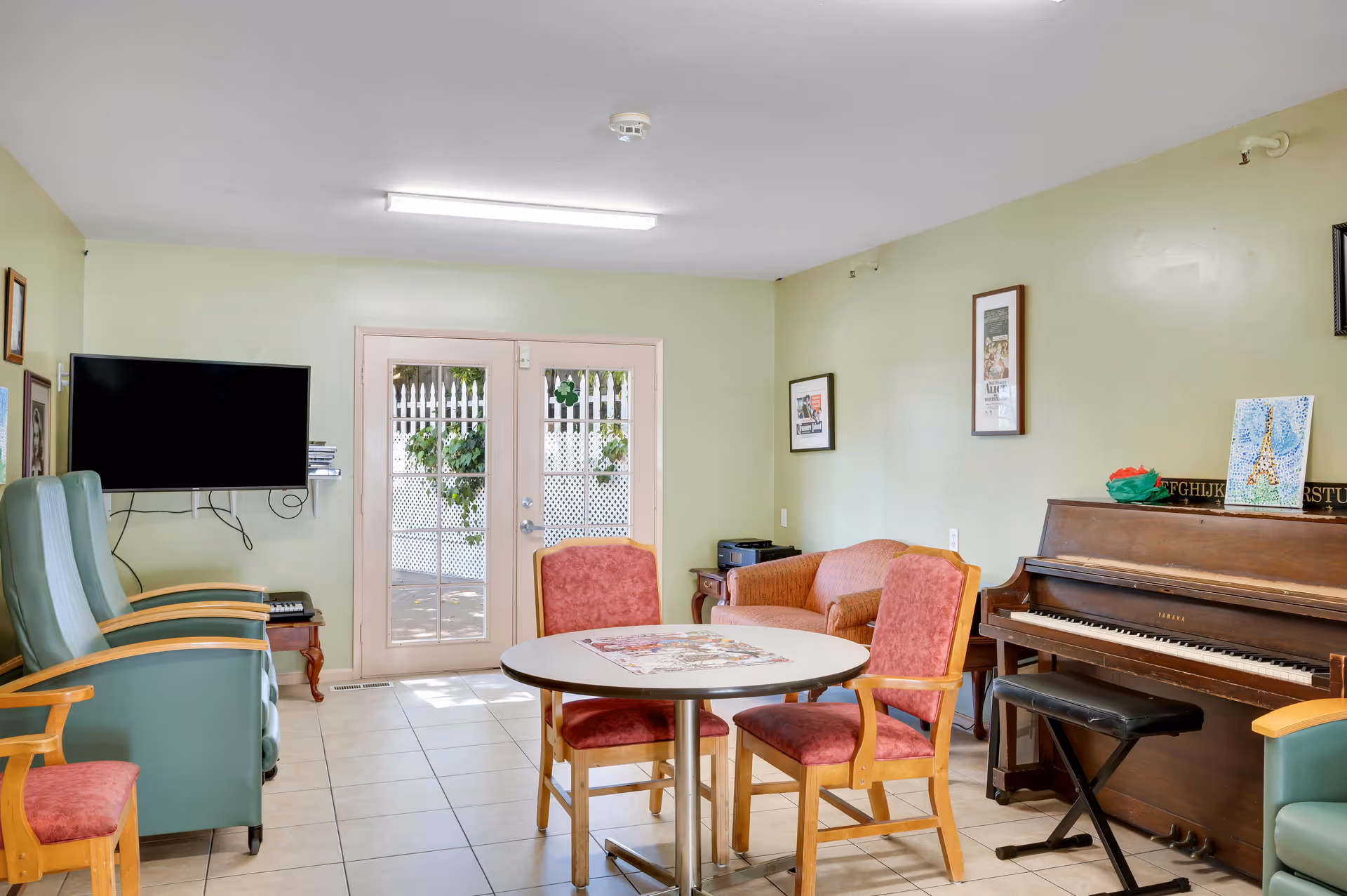 A cozy common room in Greenwood Assisted Living featuring a round table with four red cushioned chairs, a wooden piano with a black bench, a small orange couch, two green armchairs, a wall-mounted flat-screen TV, and French doors leading outside. The walls are light green with framed pictures hanging on them.