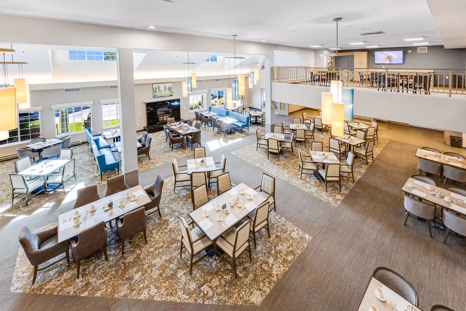 Spacious and well-lit dining area with multiple tables and chairs arranged neatly. The room features large windows allowing natural light, modern pendant lighting, a fireplace, and a mezzanine level with additional seating and a wall-mounted TV.