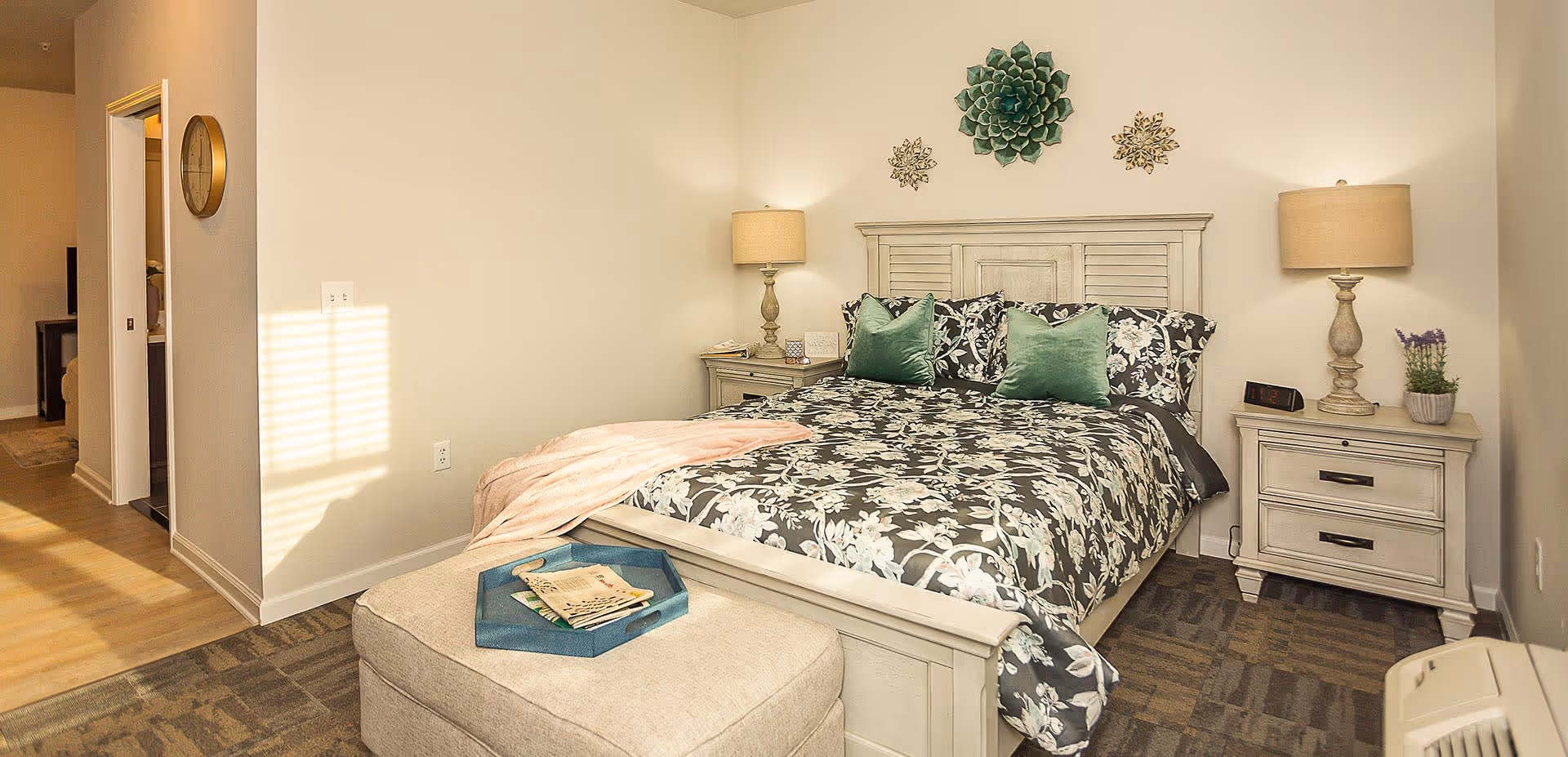 A cozy bedroom with a floral-patterned bedspread and two green pillows on a white wooden bed frame. There are two matching nightstands with lamps on either side of the bed. Above the bed, there are three decorative wall hangings. A beige ottoman with a blue tray holding some papers is placed at the foot of the bed. The room has light-colored walls and carpeted flooring, with a doorway leading to another room with wooden flooring.
