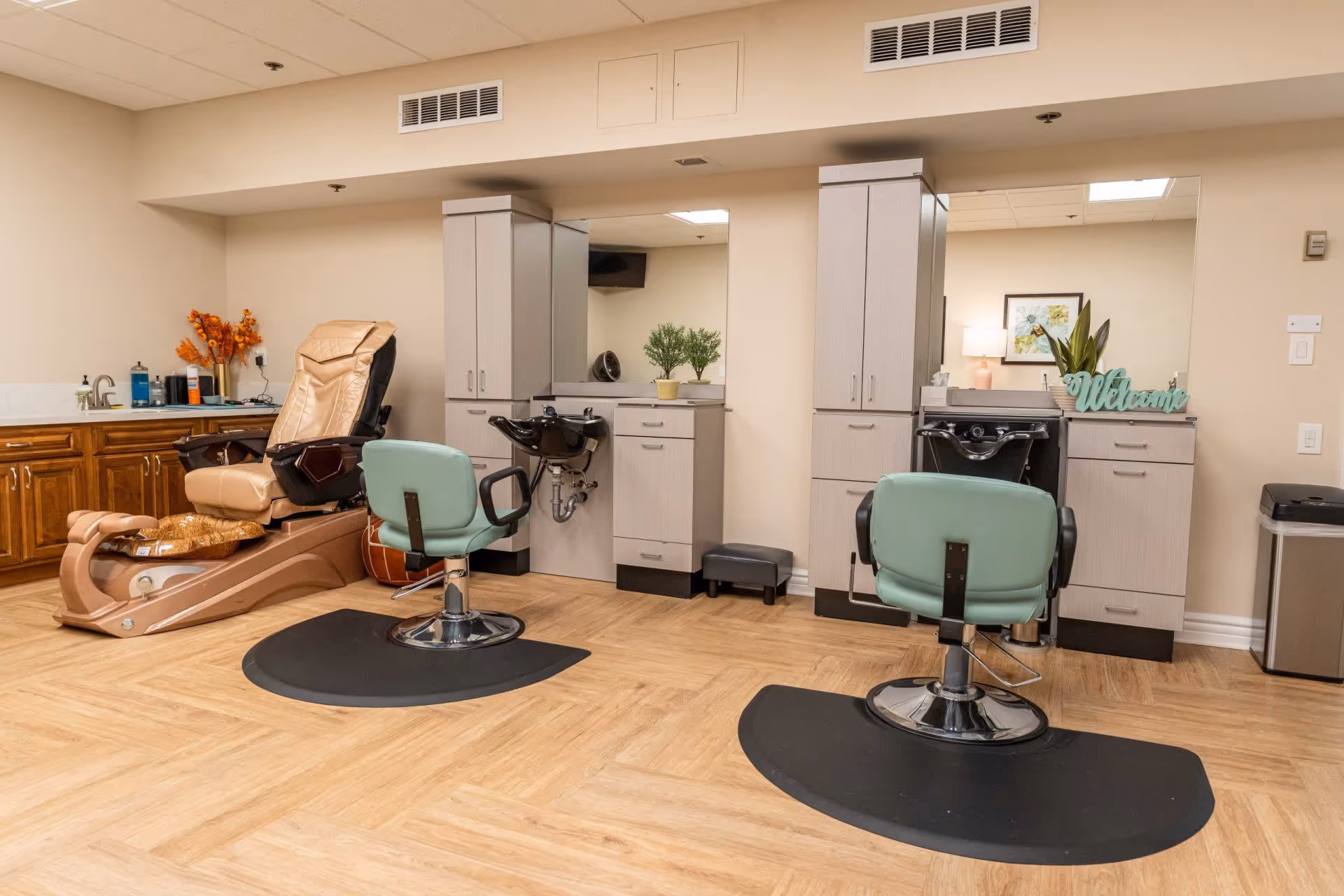 Interior view of a salon area in a senior living facility featuring two green salon chairs in front of wash basins and mirrors, a beige pedicure chair, wooden cabinets, and decorative plants and flowers.