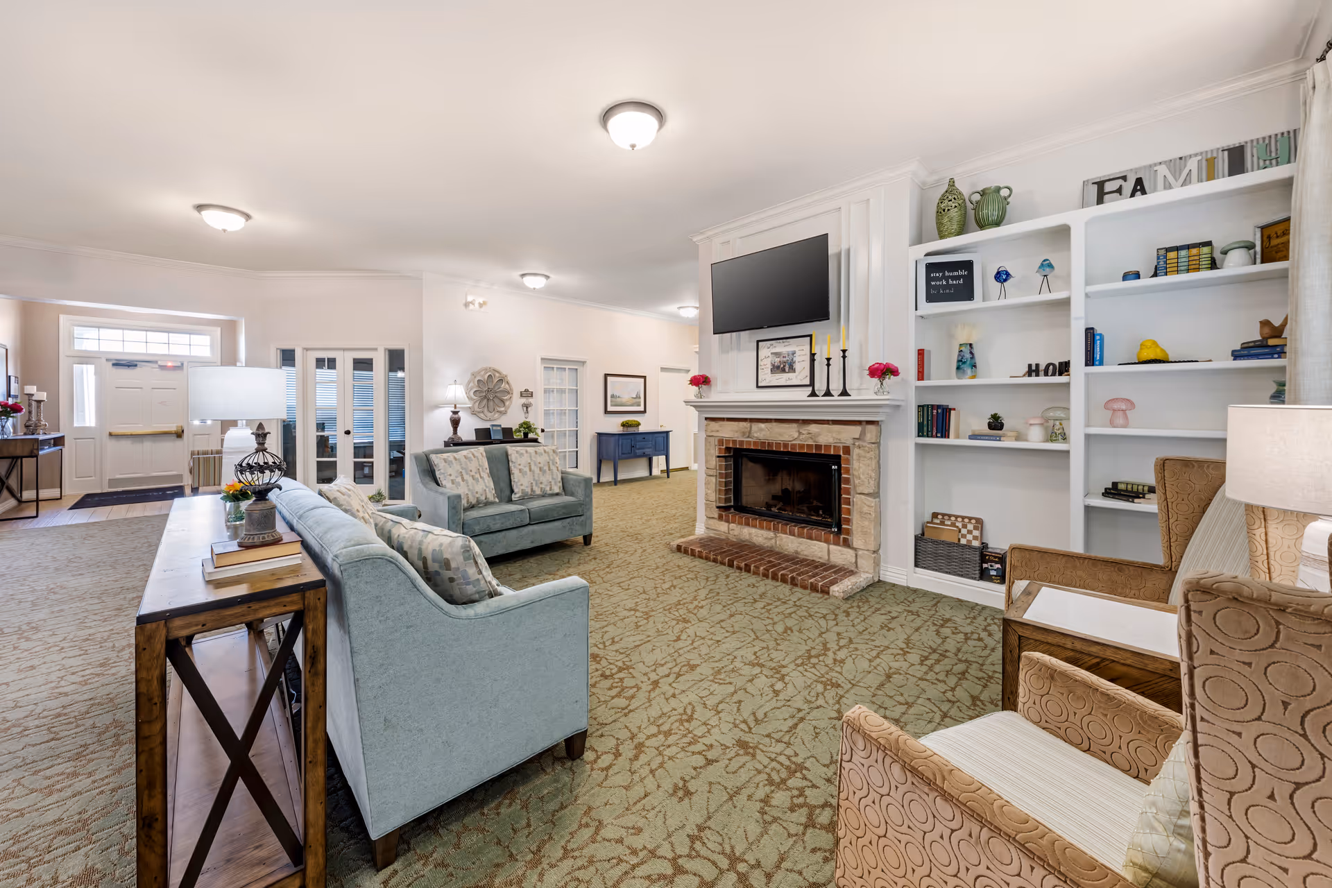 Spacious furnished common living room with sofas and armchairs facing a fireplace with a mounted TV and built-in bookshelves.