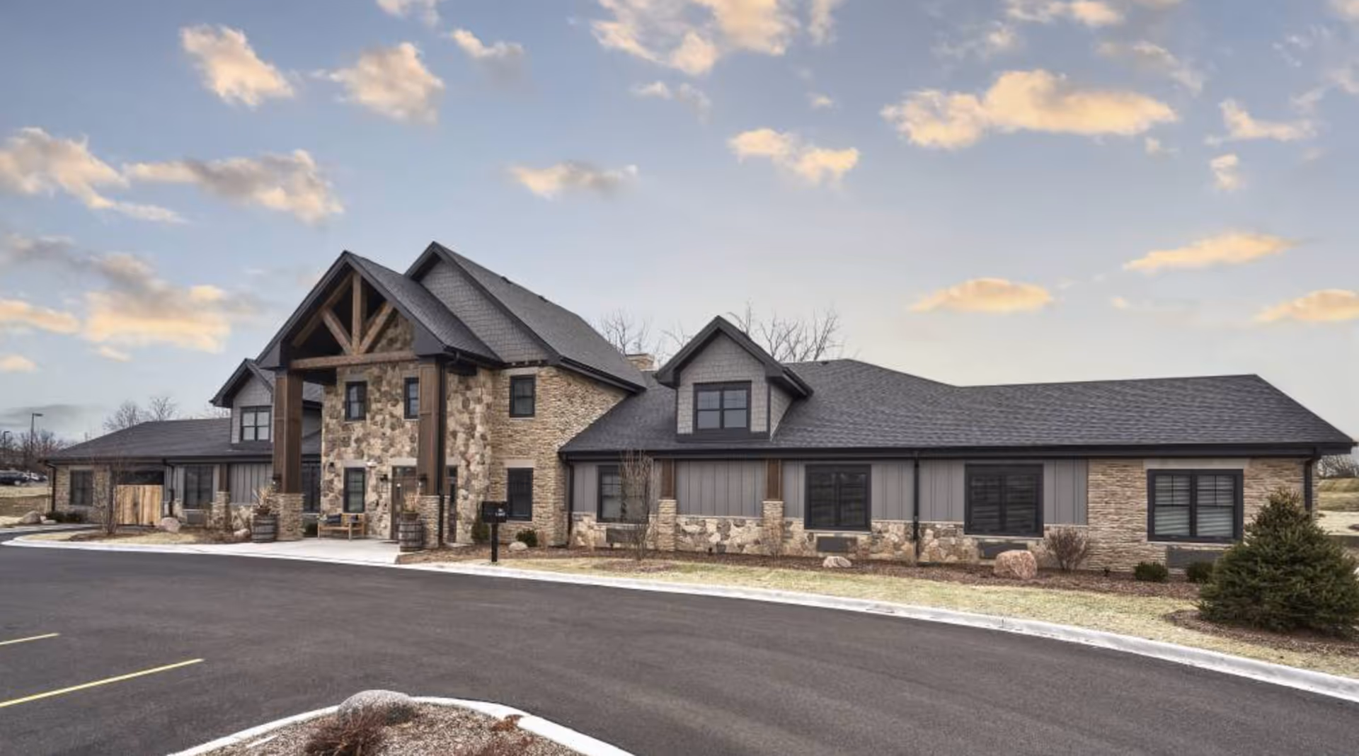 Exterior view of a single-story senior living facility building with stone and wood accents, a peaked roof, and a covered entrance. The building is surrounded by a paved driveway and some landscaping with bushes and small trees under a partly cloudy sky.