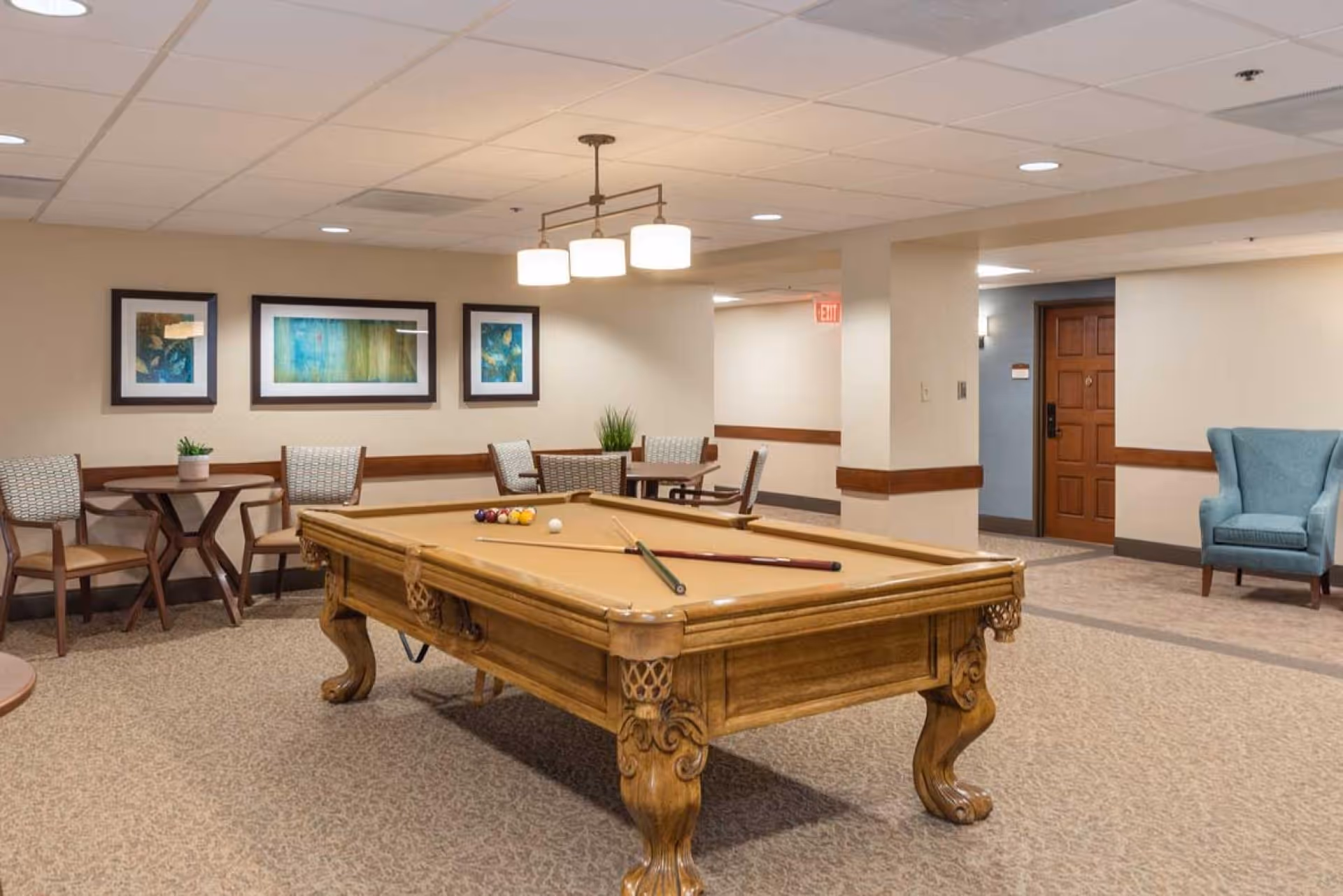 A common area in a senior living facility featuring a wooden pool table with cues and balls set up for a game. The room has carpeted floors, beige walls, and a ceiling with recessed lighting and a hanging light fixture. There are several tables and chairs along the wall, framed artwork, a blue armchair, and a wooden door in the background.