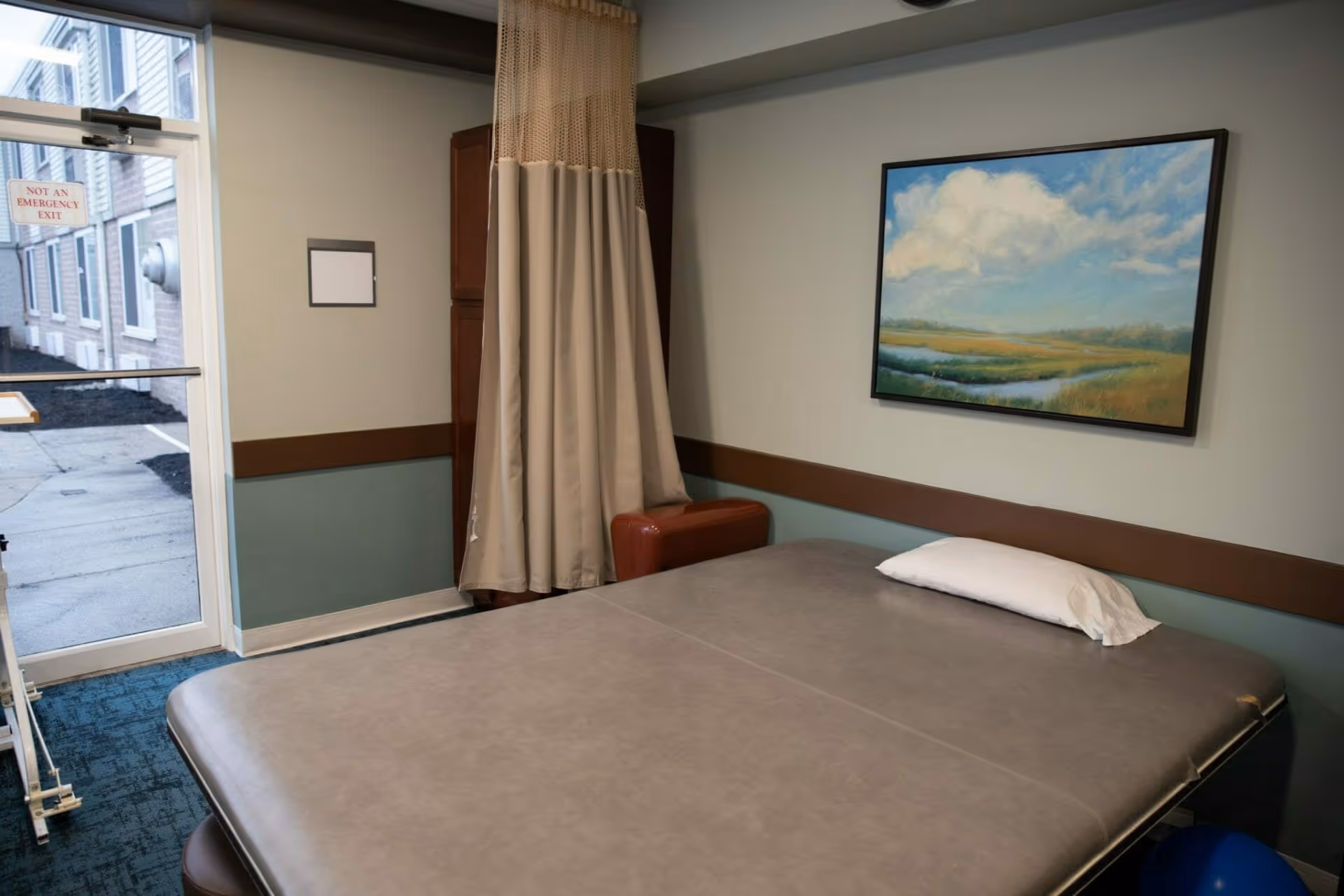 A room with a large padded examination or therapy table covered in gray vinyl with a white pillow on it. There is a beige privacy curtain partially drawn around the corner, a brown cushioned stool, and a landscape painting of a cloudy sky over a marsh hanging on the wall. A glass door with a sign that reads 'NOT AN EMERGENCY EXIT' leads outside to a paved area next to a building.