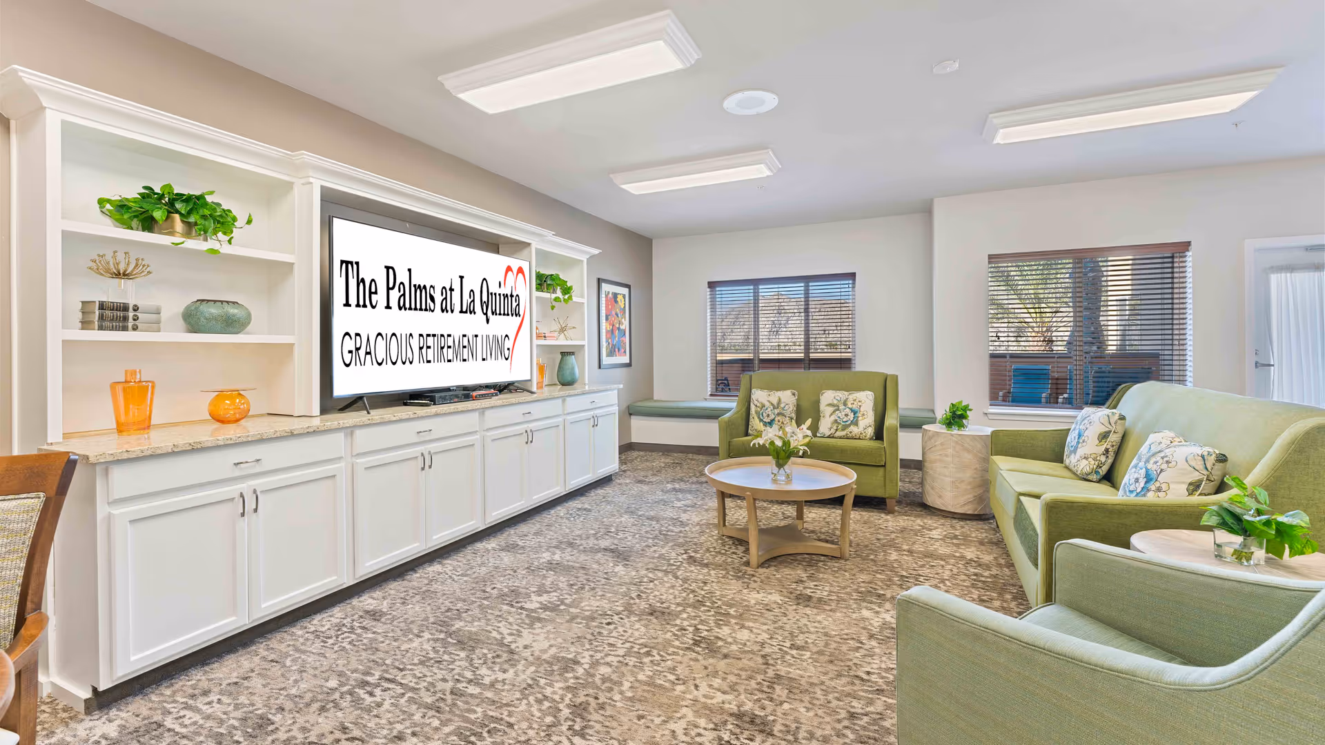 A bright and spacious living room in a retirement facility featuring green upholstered sofas and armchairs with floral cushions, a round wooden coffee table with a flower vase, built-in white cabinetry with shelves holding decorative items, and a large flat-screen TV displaying the text 'The Palms at La Quinta Gracious Retirement Living'. Two windows with blinds allow natural light into the room.