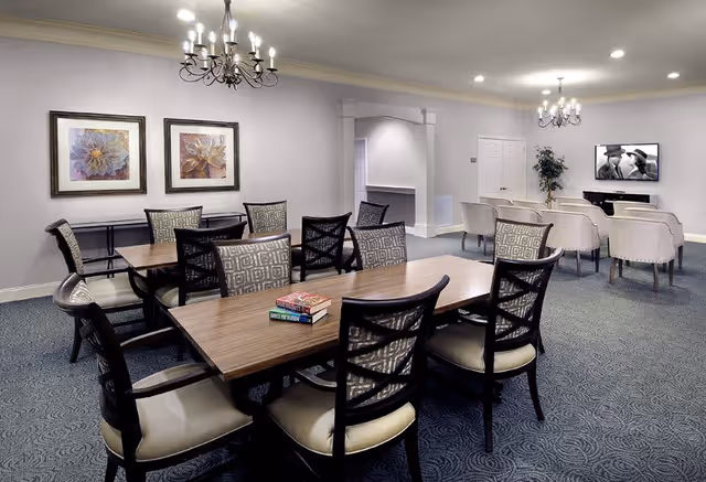 A spacious senior living common area with a large wooden table surrounded by cushioned chairs in the foreground. Two chandeliers hang from the ceiling. In the background, there are several armchairs arranged facing a wall-mounted television displaying a black and white movie scene. The room has carpeted floors, light-colored walls, and two framed floral artworks on one wall.
