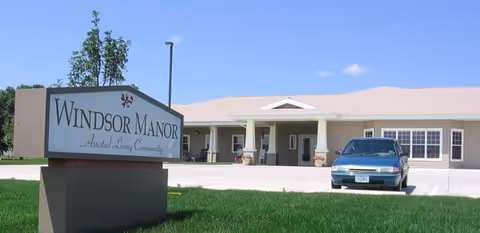 Exterior view of Windsor Manor Assisted Living Community building with a sign in the foreground and a car parked in front of the entrance on a sunny day.