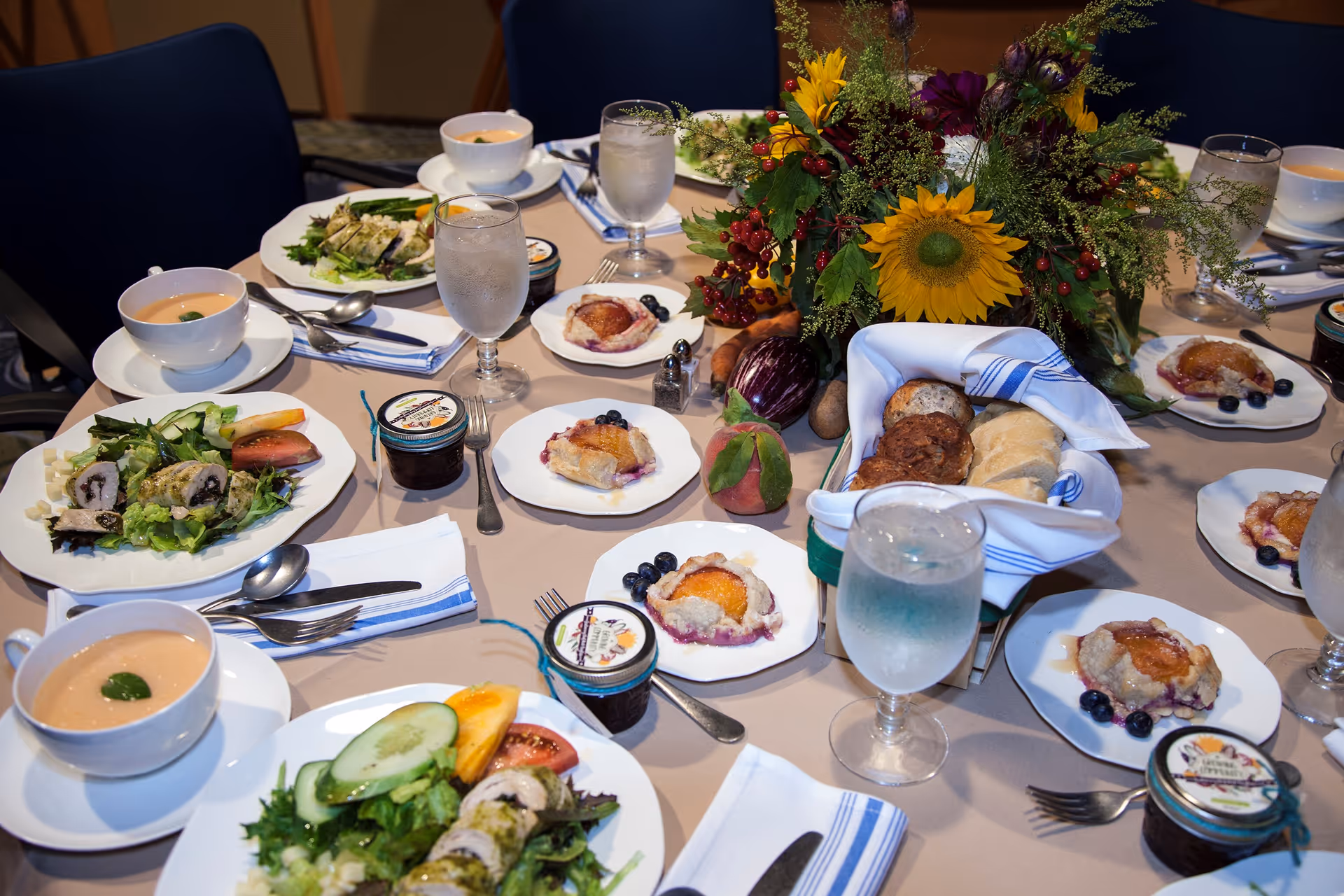 A dining table set with plates of salad, bowls of soup, glasses of water, small fruit tarts with blueberries, jars of jam, and a basket of assorted bread rolls. The table is decorated with a centerpiece of sunflowers and other greenery. Napkins and cutlery are neatly arranged around the plates.