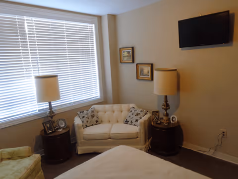 A cozy living area with a white tufted loveseat adorned with two patterned pillows, flanked by two round wooden side tables each holding a lamp and framed photos. A flat-screen TV is mounted on the beige wall above the right side table, and two small framed pictures hang on the wall above the loveseat. A large window with white blinds allows natural light into the room.