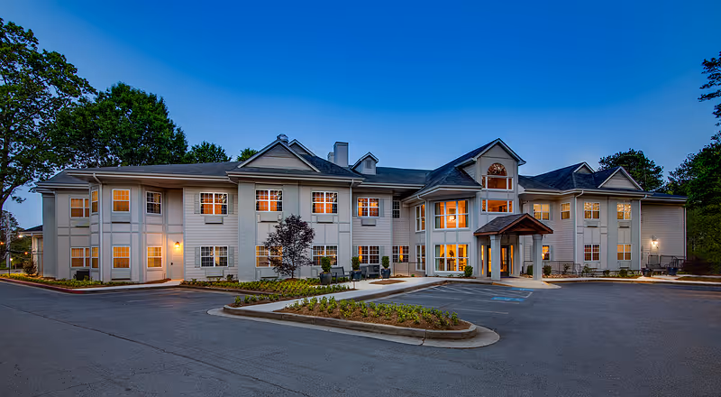 Exterior view of a two-story senior living facility building named Legacy Ridge at Buckhead during dusk, with lights glowing warmly from the windows and a clear blue sky overhead. The building has a welcoming entrance with a covered porch and surrounding landscaping, including trees and shrubs, and a paved parking area in front.