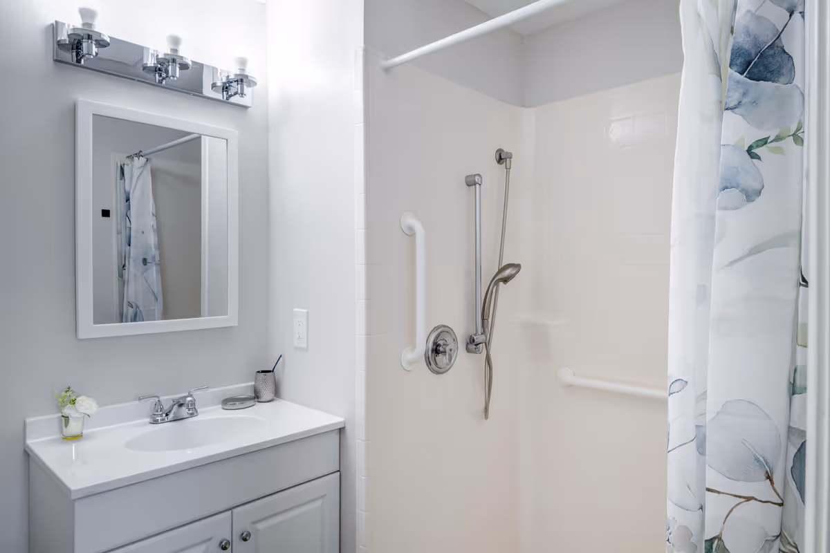 A clean bathroom featuring a white vanity with a sink, a wall-mounted mirror with three light fixtures above it, and a shower area with white tiled walls, grab bars, a handheld showerhead, and a floral-patterned shower curtain.