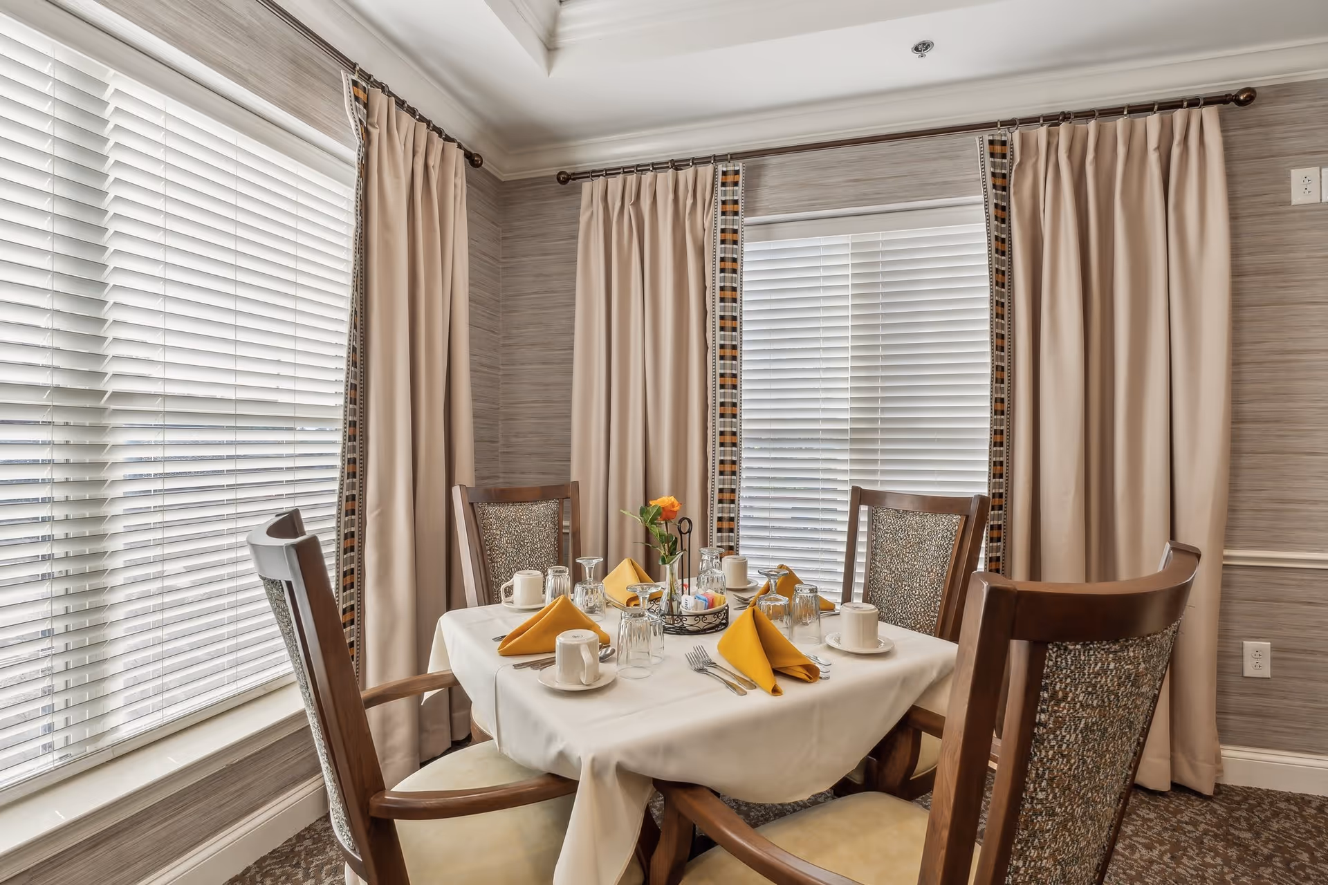 A small dining table set for four with yellow napkins, cups, and place settings in a sunlit room with large windows and beige curtains.