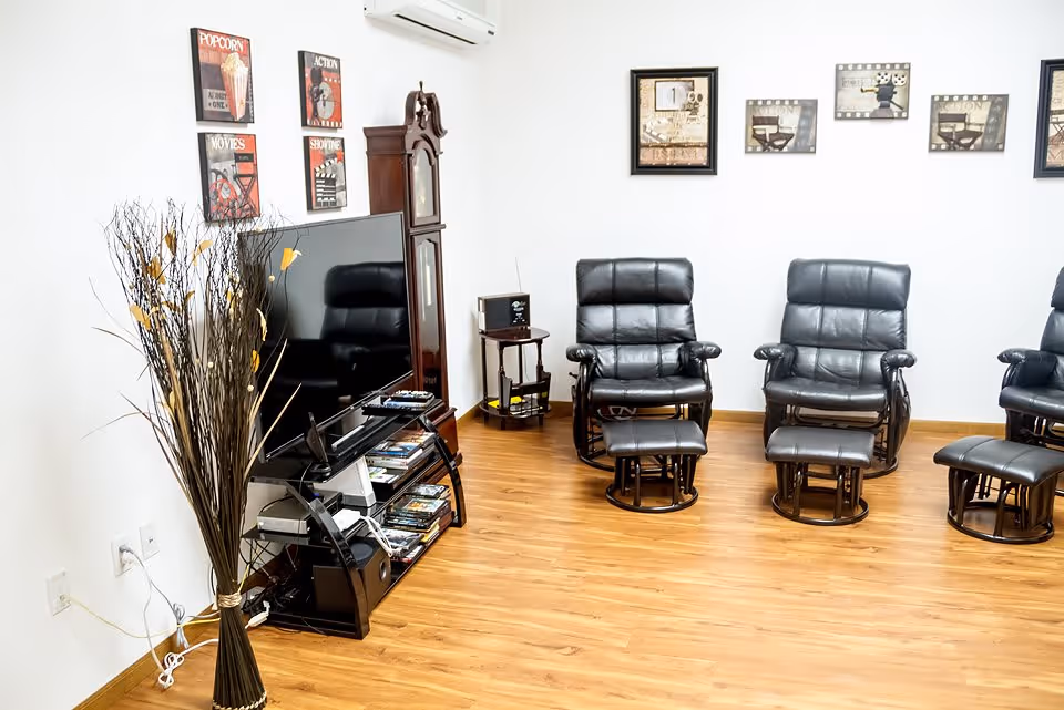 A cozy living room area with three black leather recliner chairs and matching footrests arranged on a wooden floor. A large flat-screen TV is placed on a black TV stand with shelves holding DVDs and electronic devices. A tall grandfather clock stands next to the TV. Decorative wall art with movie themes and vintage film motifs adorn the white walls. A tall vase with dried branches is positioned near the TV stand.