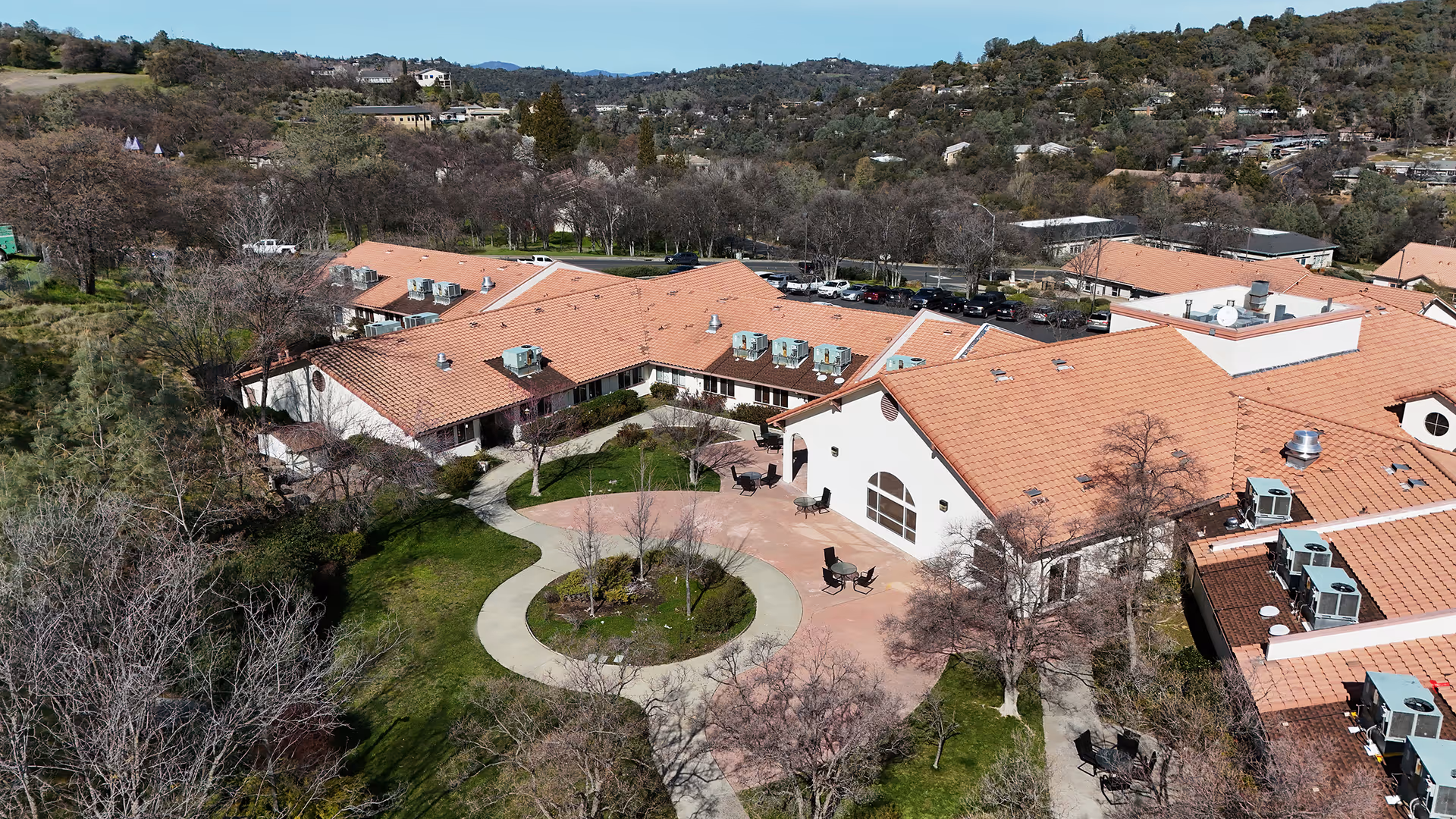 Aerial view of Sonora Convalescent Hospital showing a large building with terracotta roof tiles surrounding a circular courtyard with a paved walkway and some outdoor seating. The building is set in a green landscape with trees and hills in the background under a clear blue sky.