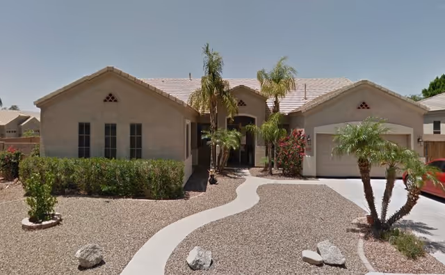 Front exterior view of a single-story assisted living facility named Mountain Vista Manor Corp Assisted Living, featuring a beige stucco facade, a tiled roof, a curved concrete walkway, desert landscaping with gravel, palm trees, and shrubs.
