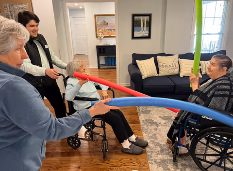 Four people in a living room setting, two elderly women in wheelchairs and two other individuals standing, engaging in a playful activity using colorful foam noodles. The room has wooden floors, a blue couch with pillows, framed artwork on the walls, and a rug.