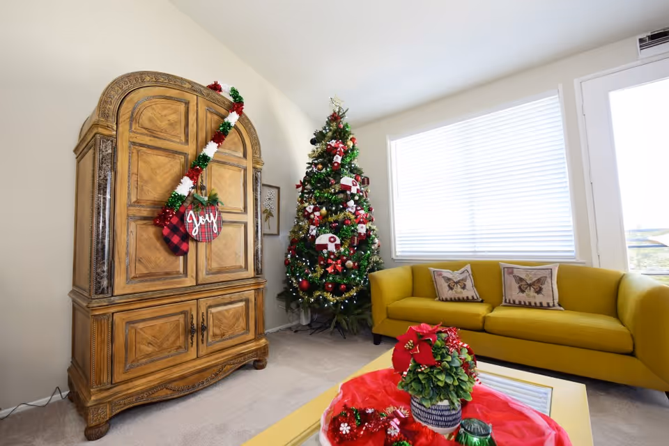 A cozy living room decorated for Christmas with a large decorated Christmas tree, a wooden cabinet adorned with a festive garland and a sign that says 'Joy', a mustard yellow sofa with butterfly-themed pillows, and a coffee table with holiday decorations including a small plant and red fabric.