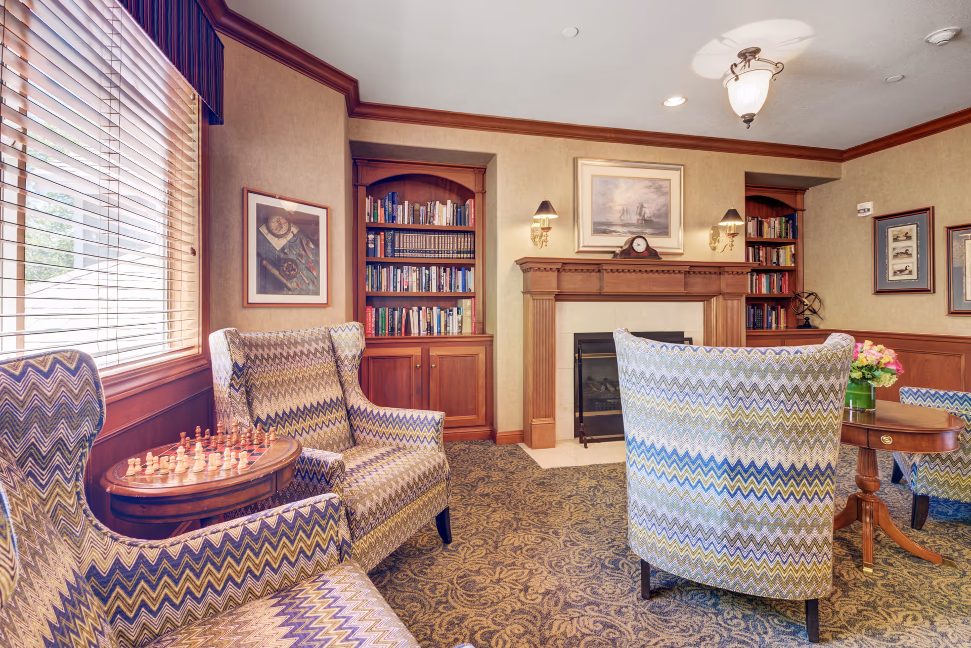 A cozy living room area with patterned armchairs arranged around a wooden coffee table and a fireplace. Built-in wooden bookshelves flank the fireplace, filled with books. The room features a large window with blinds, framed artwork on the walls, and a carpeted floor with an intricate design.
