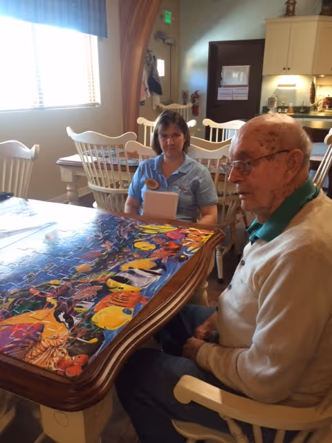 An elderly man and a woman sitting at a wooden table working on a colorful jigsaw puzzle in a well-lit room with white chairs and a kitchen area in the background.