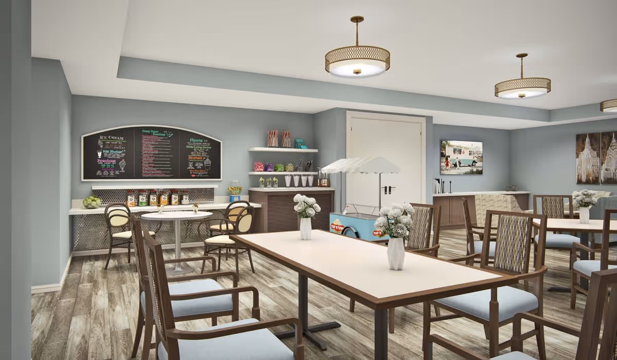 A bright and clean dining area with multiple tables and chairs, decorated with small vases of white flowers. The room has light blue walls and wood-patterned flooring. On one side, there is a counter with jars of snacks and a menu board above it, along with shelves holding candy and cups. There is also a small cart with an umbrella, likely for serving ice cream or treats. The room is well-lit with modern ceiling lights and has artwork on the walls.