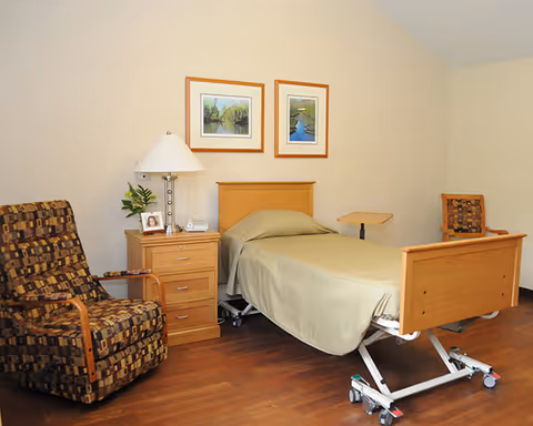 A simple, clean bedroom in a senior living facility featuring a single adjustable hospital-style bed with beige bedding. Next to the bed is a wooden nightstand with a lamp, a small plant, a framed photo, and a telephone. There are two framed landscape paintings on the beige wall above the bed. The room also includes two patterned armchairs with wooden armrests and a small wooden side table. The floor is hardwood.