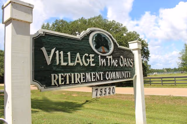 Wooden sign for Village In The Oaks Retirement Community with the number 75520, set outdoors with grass, trees, and a partly cloudy sky in the background.
