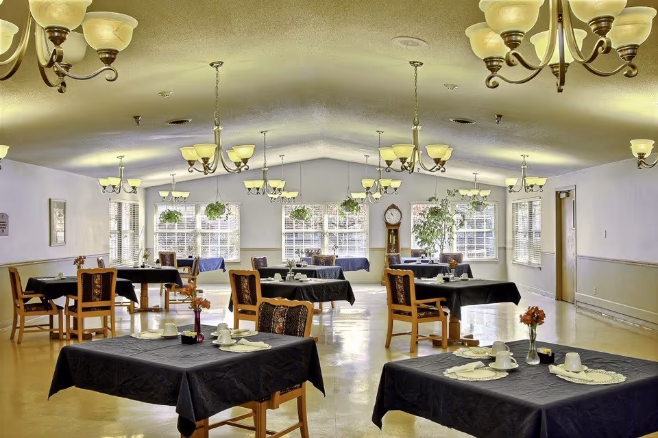 A spacious dining room with multiple tables covered in black tablecloths, each set with white cups, saucers, and napkins. Wooden chairs with cushioned seats surround the tables. The room is well-lit with multiple chandeliers hanging from the ceiling and large windows letting in natural light. There are several hanging plants near the windows and a tall grandfather clock against the far wall.
