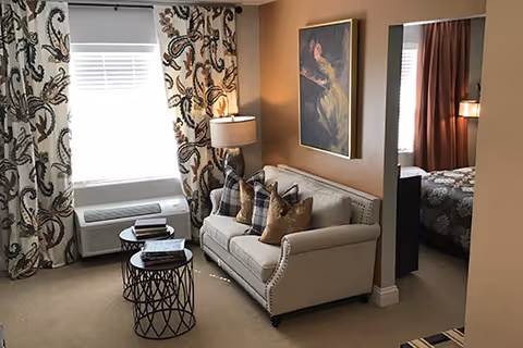 A cozy living room area with a beige sofa adorned with multiple pillows, a floor lamp beside it, and two round nested coffee tables with books on top. The room has patterned curtains framing a window with blinds, and a framed painting hangs on the wall above the sofa. An adjacent bedroom with a bed and curtains is visible through an open doorway.