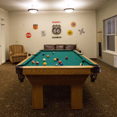 A recreational room with a pool table in the center, a brown leather couch against the back wall, a striped armchair to the left, and a rack of pool cues to the right near a window. The wall behind the couch is decorated with various vintage road signs including Route 66, Stop, and No Speed Limit signs.