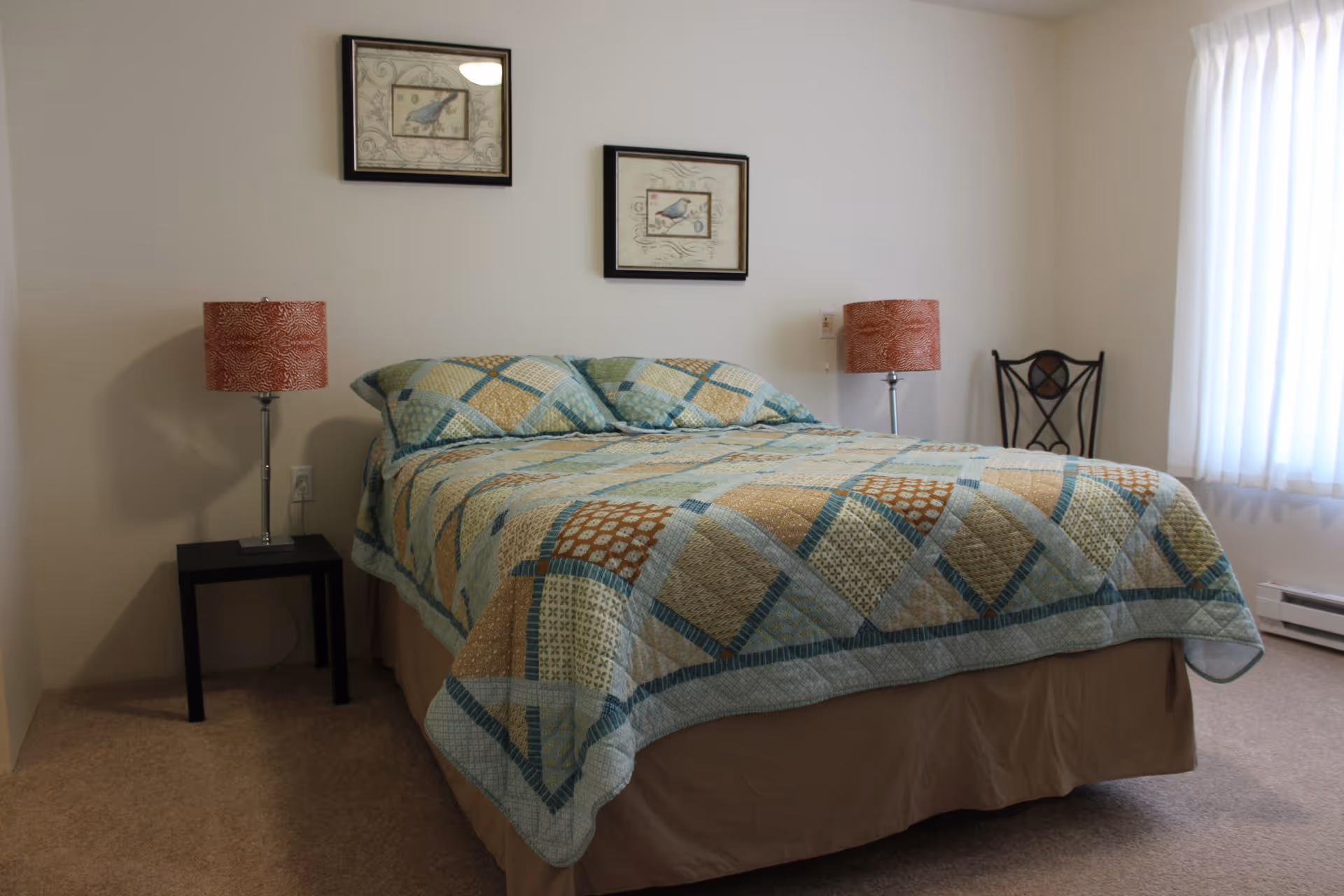 A bedroom with a neatly made bed featuring a patterned quilt in shades of blue, green, and beige. There are two matching pillows on the bed. On either side of the bed are small black nightstands, each with a red patterned lamp. Two framed pictures of birds hang on the wall above the bed. A window with white curtains allows natural light into the room, and a decorative chair is placed near the window.