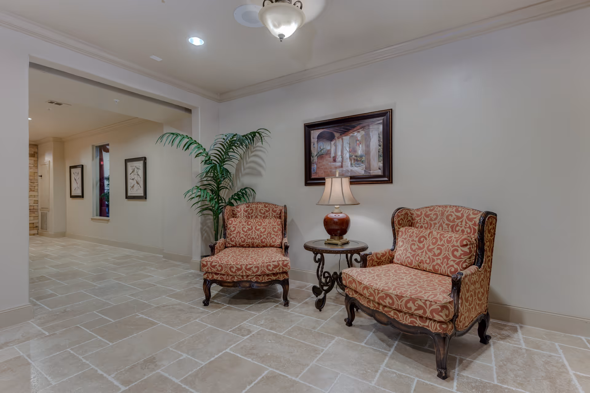 A seating area in a hallway with two upholstered armchairs featuring red and gold patterned fabric, a small round table with a lamp between them, a potted plant behind one chair, and a framed painting on the wall above the table. The floor is tiled with light-colored stone, and the walls are painted a light neutral color.