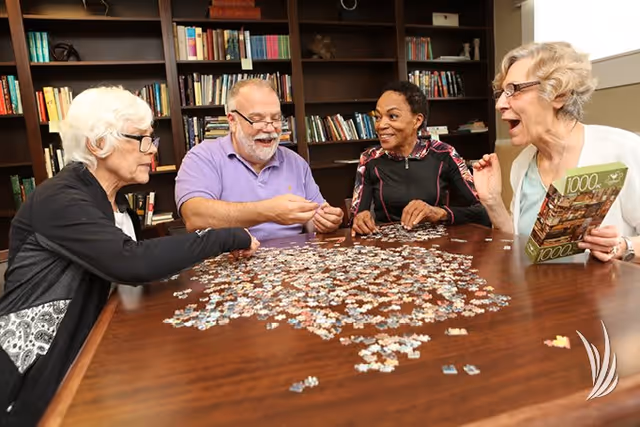 Four elderly adults sitting around a wooden table working together on assembling a large jigsaw puzzle in a room with bookshelves filled with books in the background. One woman is holding the puzzle box, and all appear engaged and happy.