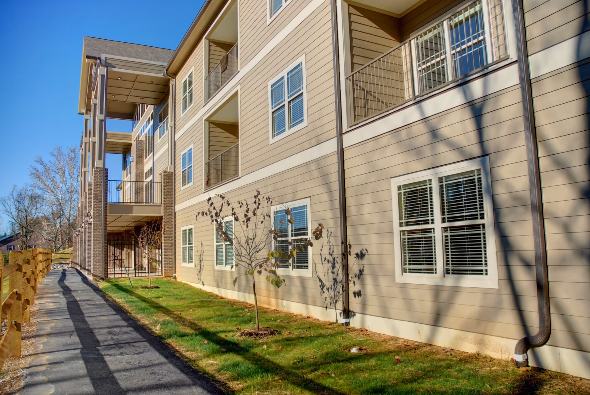 Exterior view of a multi-story residential building with beige siding and white-framed windows. There is a paved walkway alongside the building, bordered by a wooden fence and small trees planted in a grassy area. The sky is clear and blue.