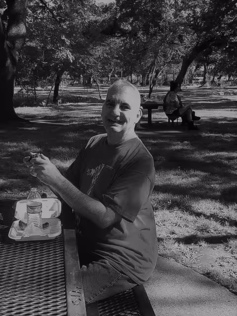 A man sitting at a picnic table in a park, smiling at the camera. He has a water bottle and a food container on the table. In the background, another person is sitting on a bench under the shade of trees.