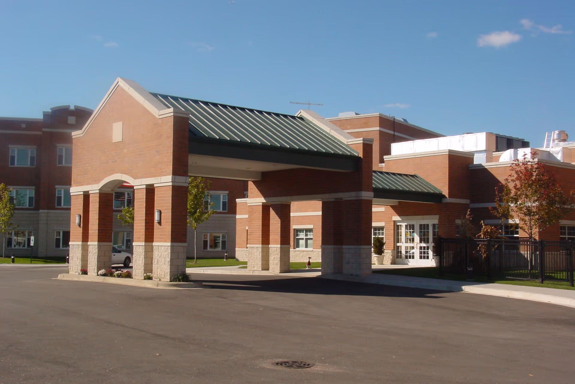 Exterior view of a senior living facility building with a covered entrance supported by brick columns and a green metal roof. The building is made of red brick with beige stone accents and has multiple windows. There are small trees and a clear blue sky in the background.