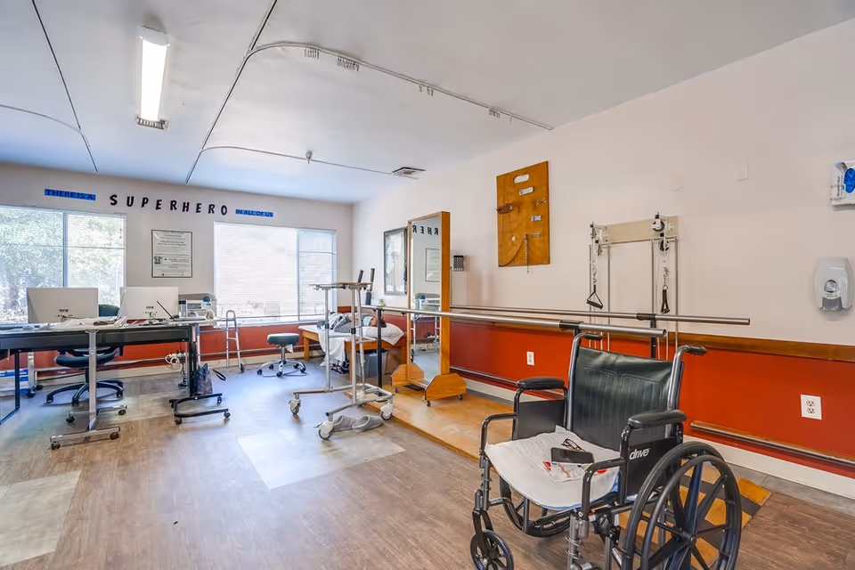 A rehabilitation or physical therapy room with a wheelchair in the foreground, parallel bars for walking exercises, a mirror, and various therapy equipment. There are two desks with computers near large windows letting in natural light. The wall has a motivational phrase 'There is a superhero in all of us.'
