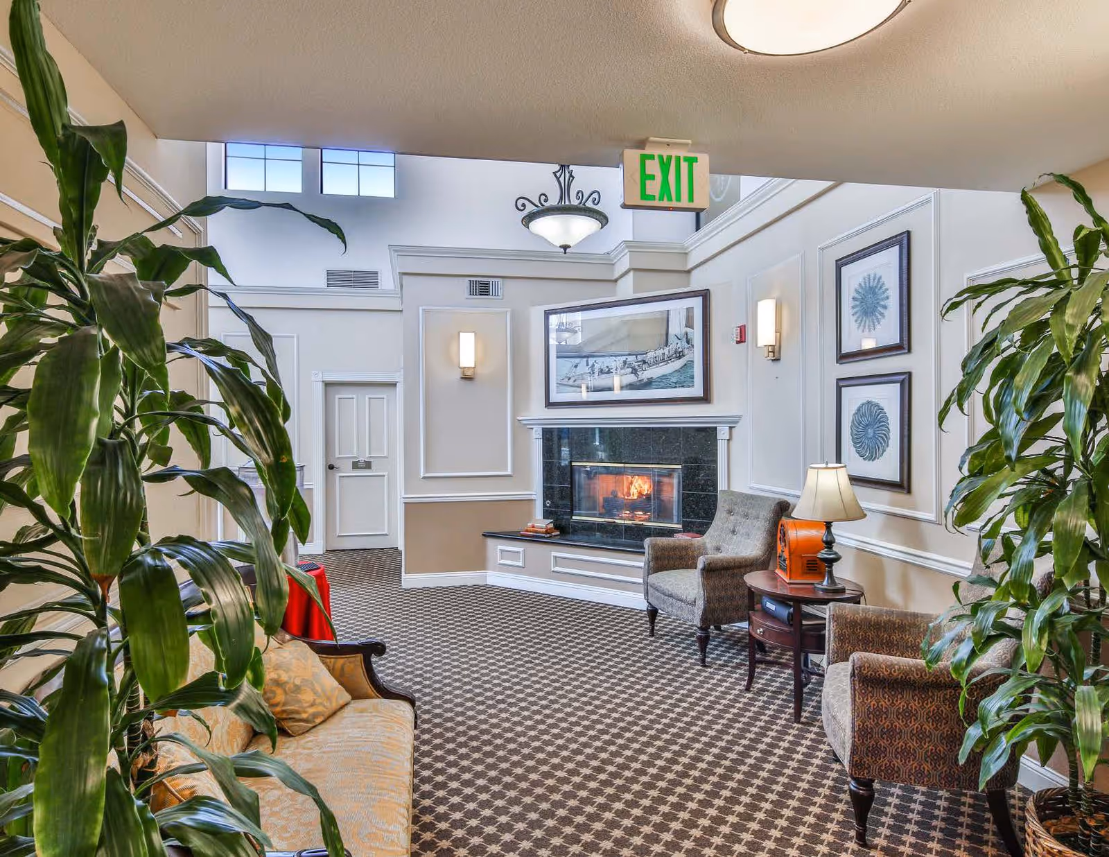 A cozy interior common area with a lit fireplace, two upholstered armchairs, a small wooden side table with a lamp, a patterned carpet, framed artwork on the walls, and large green plants on either side. There is an exit sign above a doorway in the background.